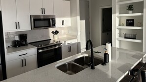 Een moderne keuken met witte kasten, roestvrijstalen apparatuur, een dubbele spoelbak en een aanrecht met een koffiezetapparaat en keukengerei.