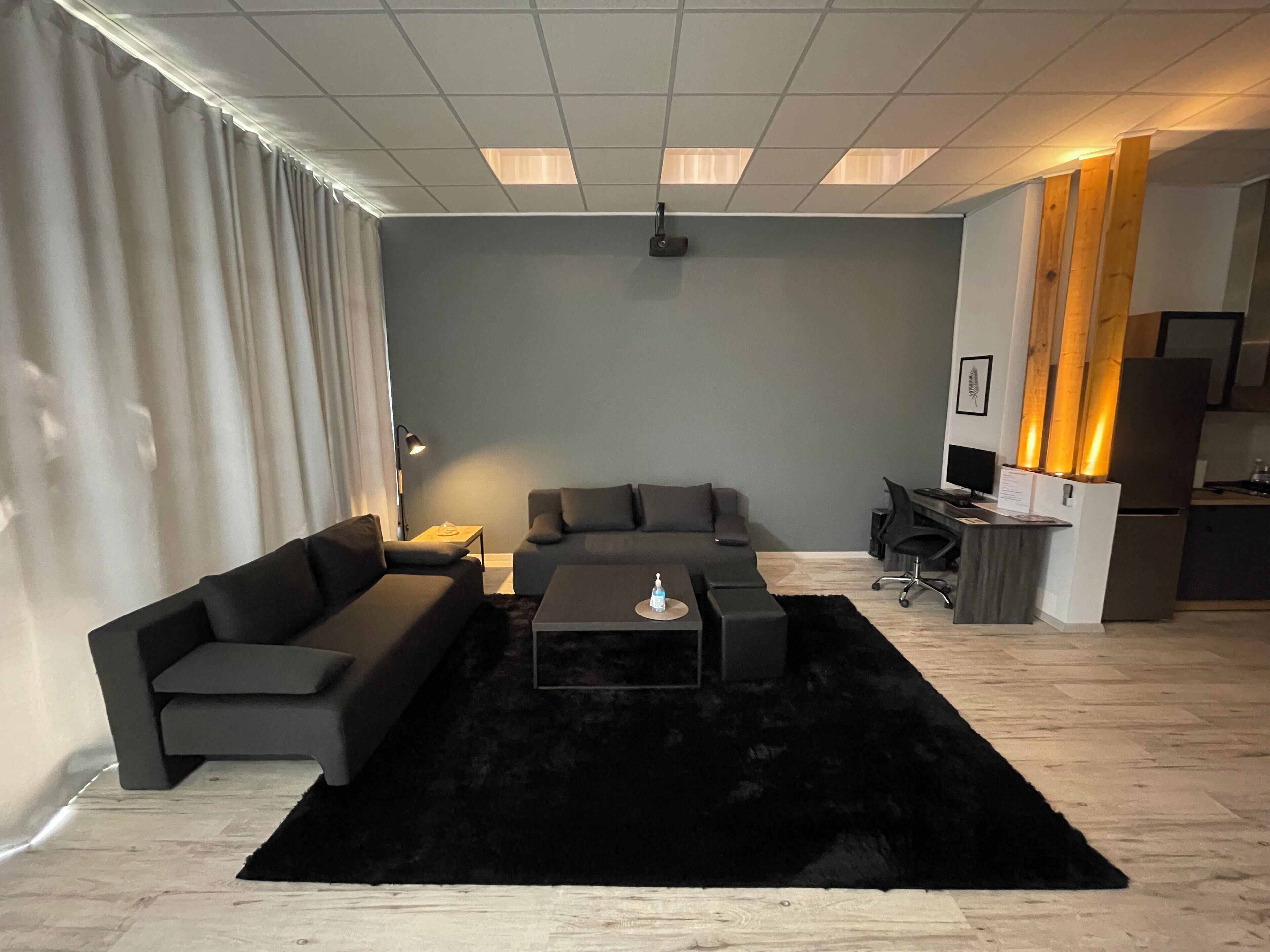 A modern living room with a grey sofa set, a black coffee table, and a black rug. There is a desk with a computer and a chair, and a kitchen area with a refrigerator and a stove.