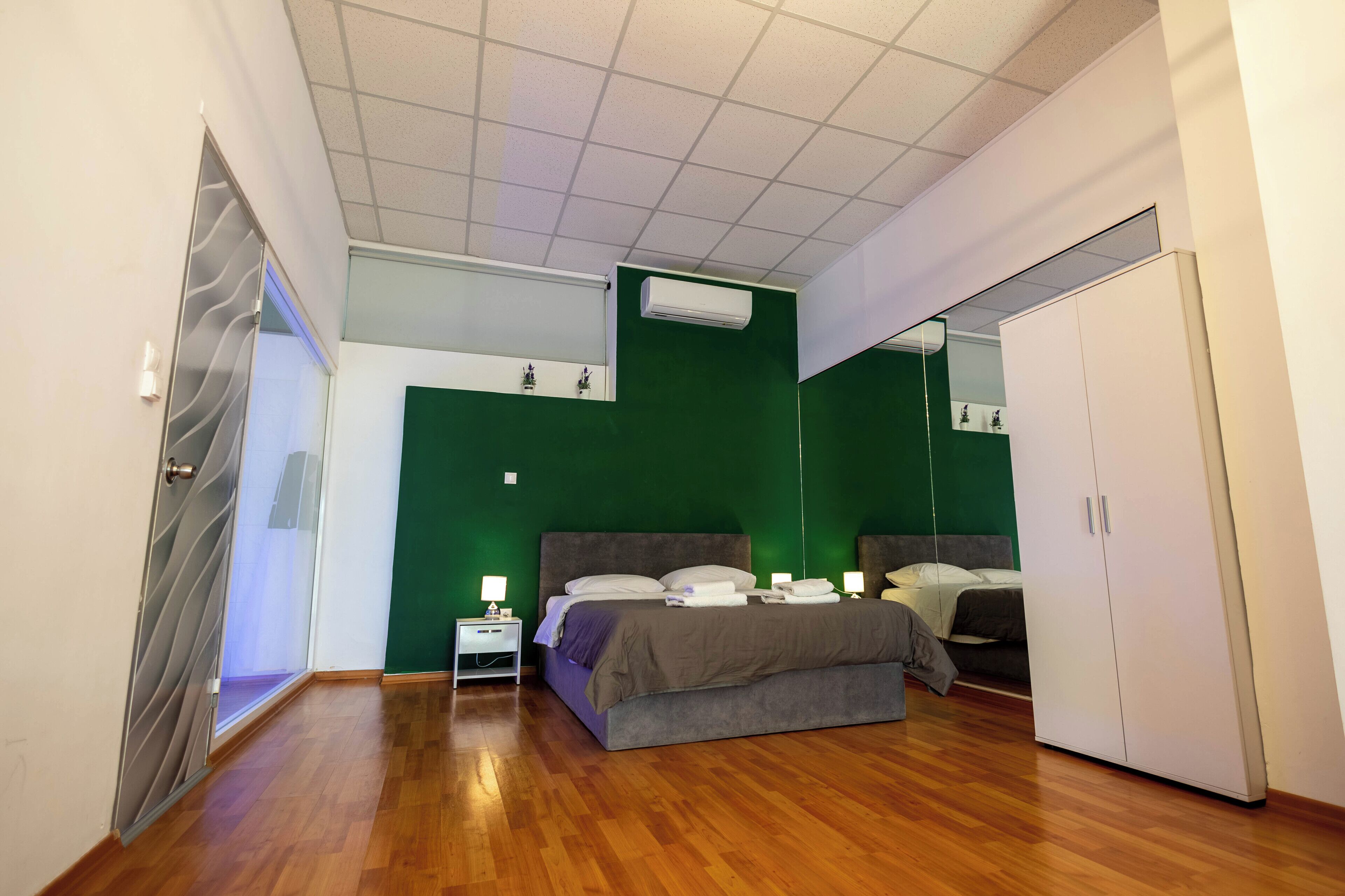 A bedroom with a green accent wall, a grey bed, a white wardrobe, and a small bedside table with a lamp.
