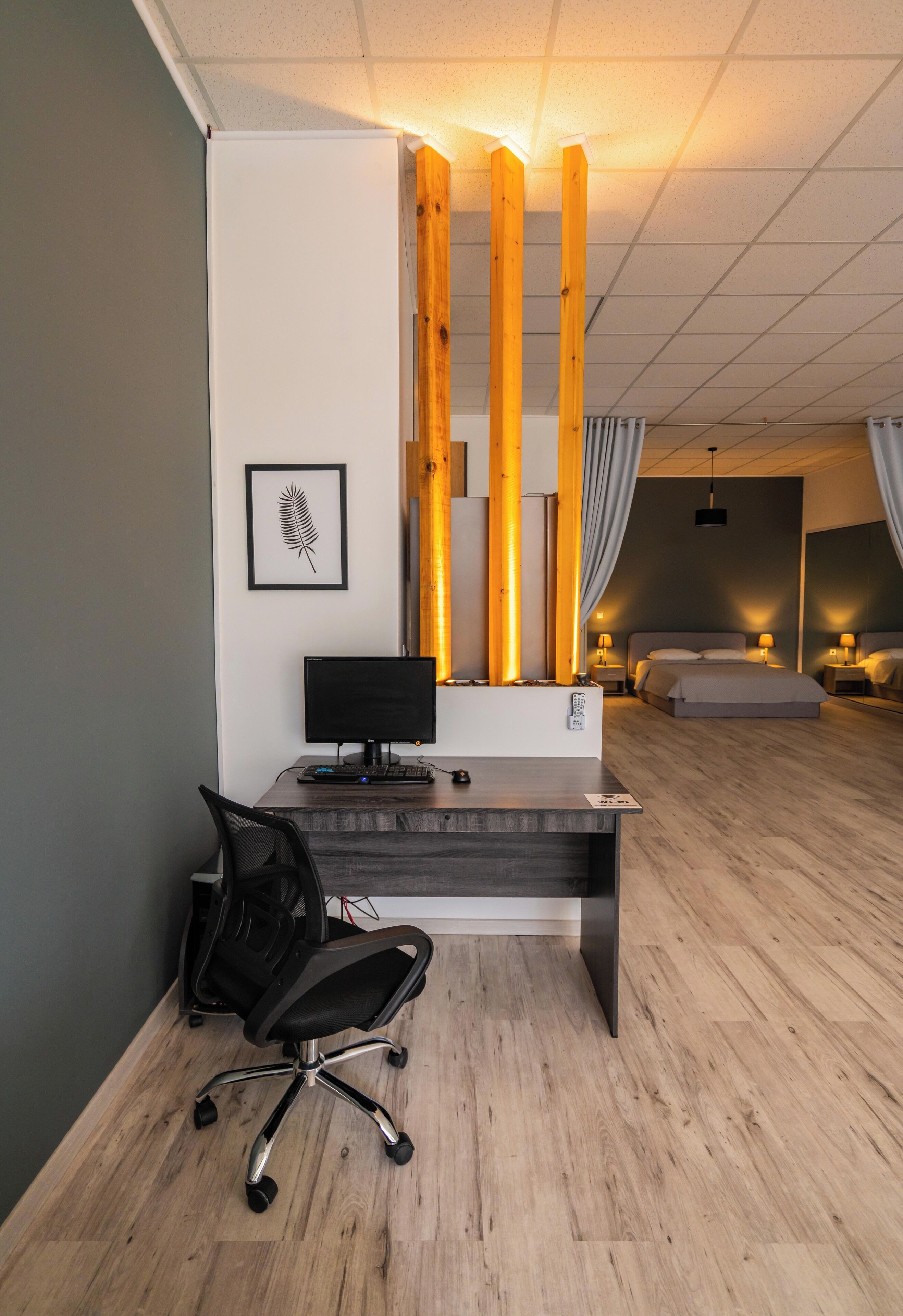 A modern hotel room with a wooden desk, a computer, a chair, a bed, and a bedside table with lamps.