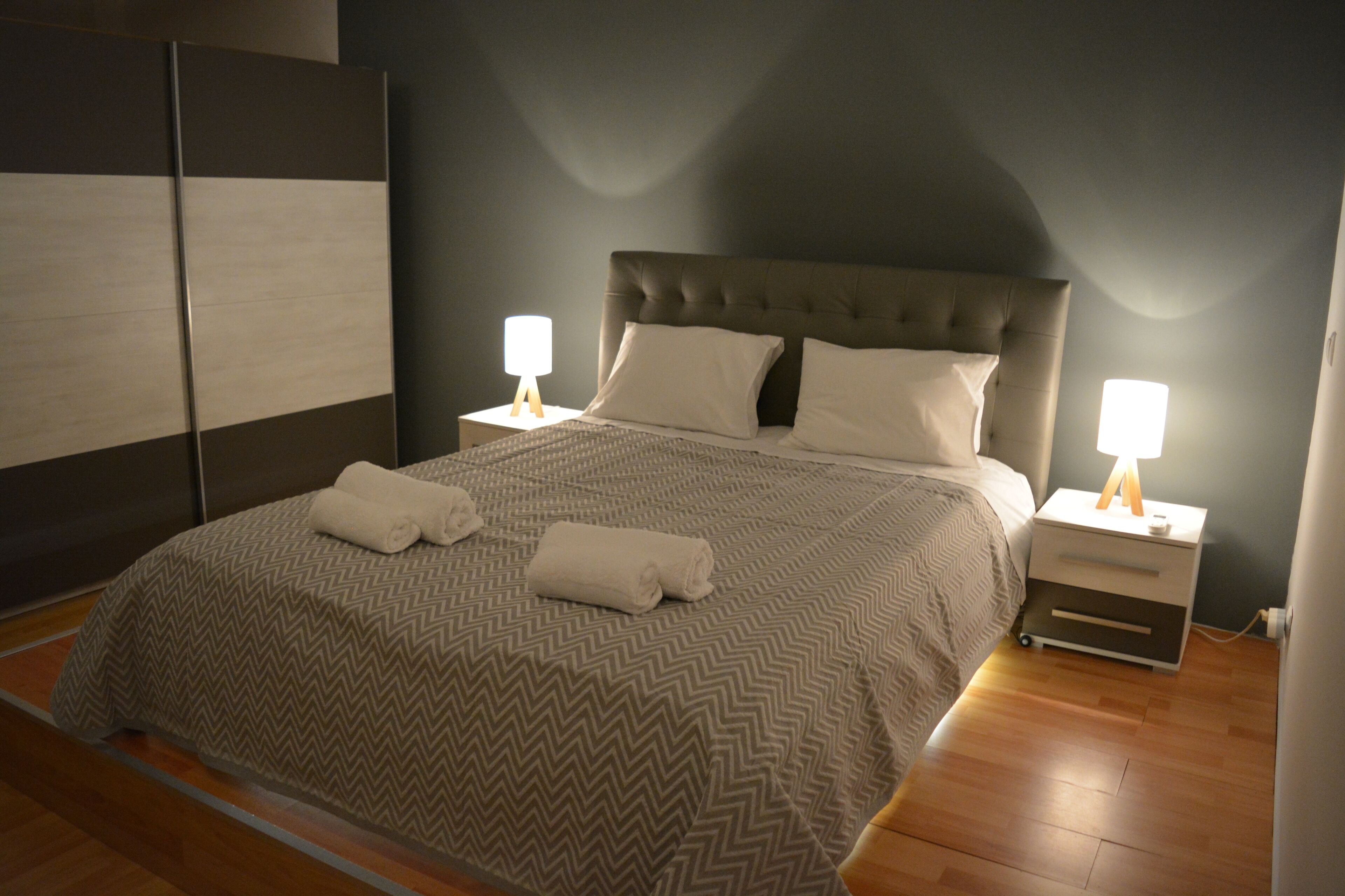 A bedroom with a large bed, two bedside tables with lamps, a wardrobe, and a wooden floor.