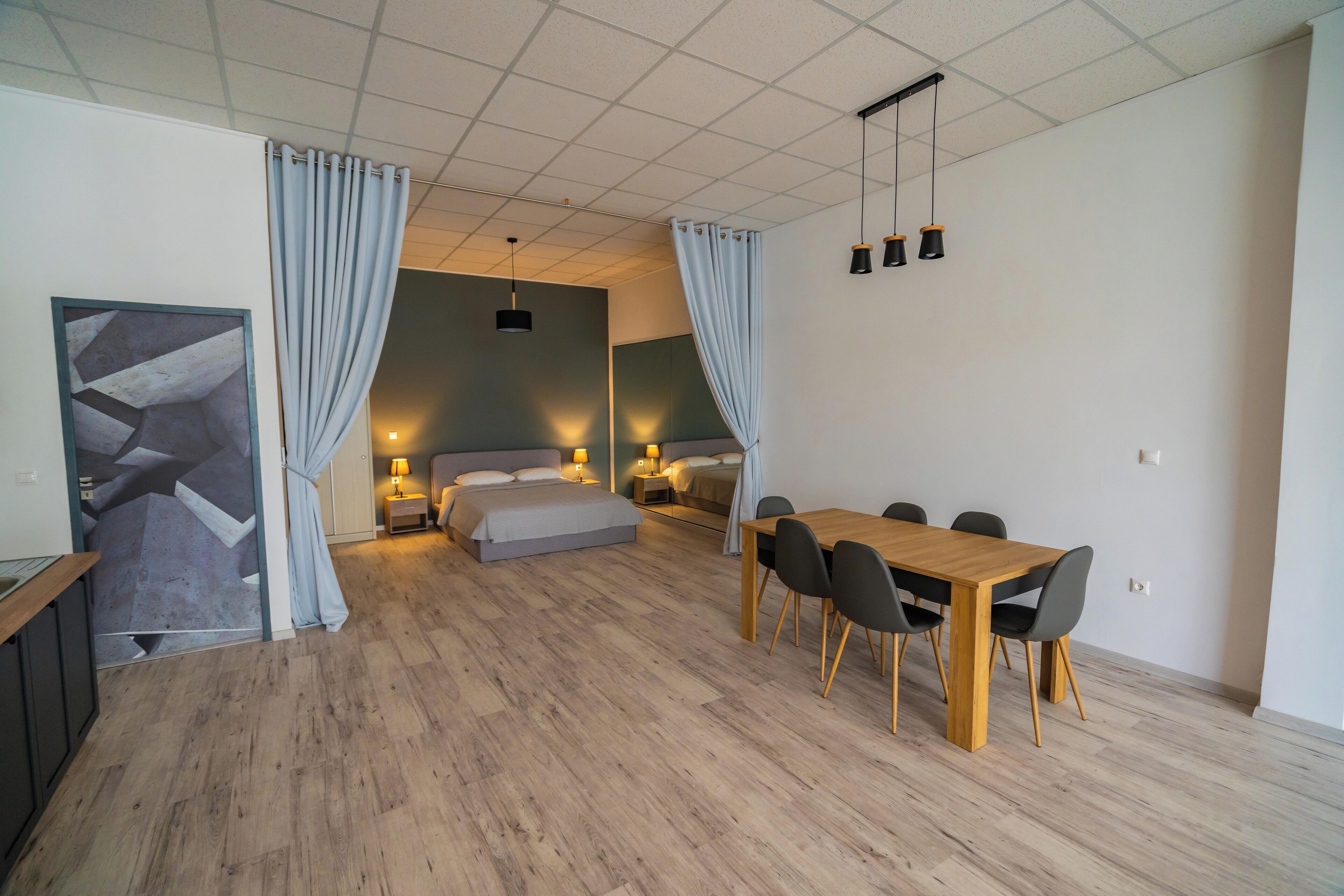 Ein modernes Schlafzimmer mit Holzboden, einem Bett, einem Nachttisch mit Lampe und einem großen Fenster mit Vorhängen.