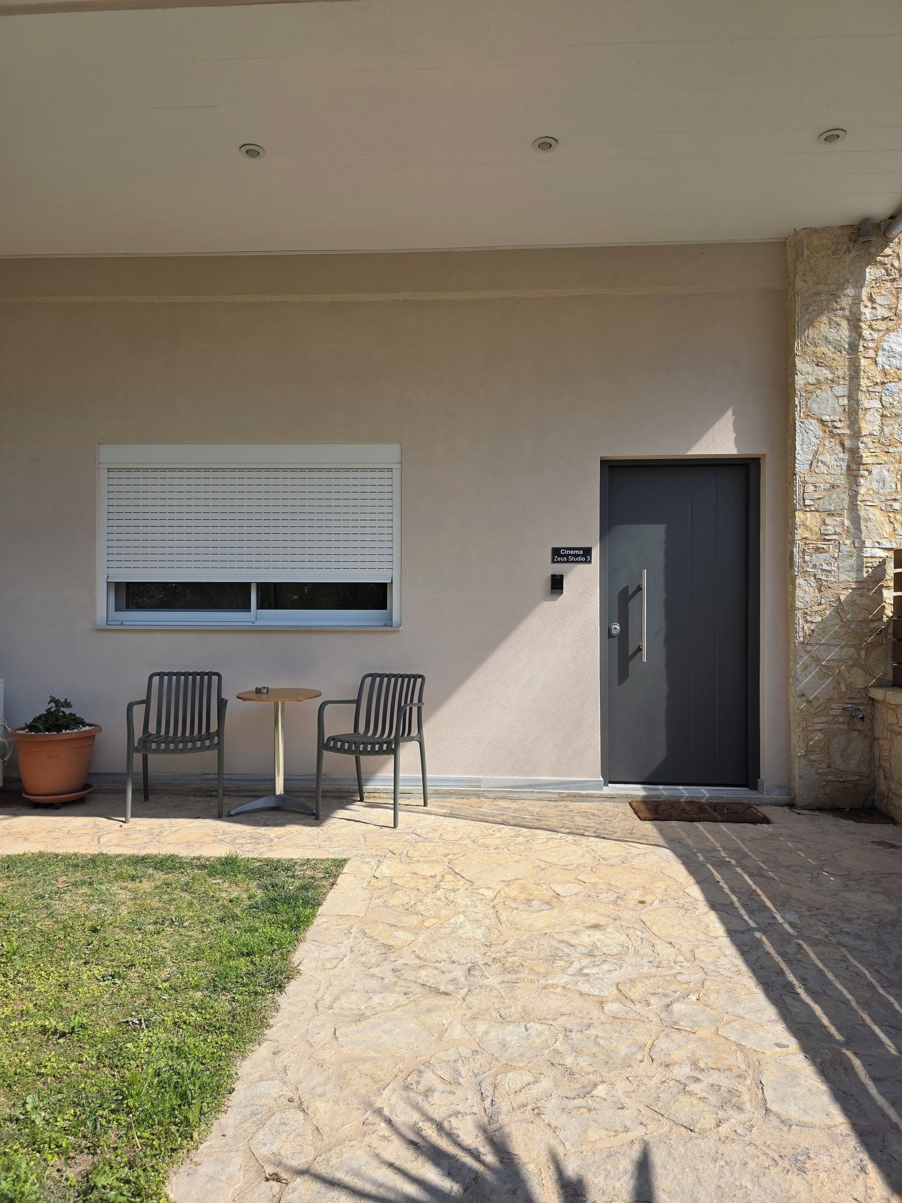 A modern building exterior with a stone wall, a door labeled "Cinema Zeus Studio 3," and two chairs outside.