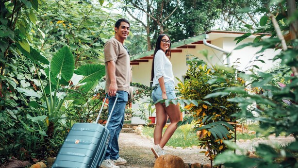 A man and a woman standing in a garden with a suitcase, surrounded by greenery and a building in the background.