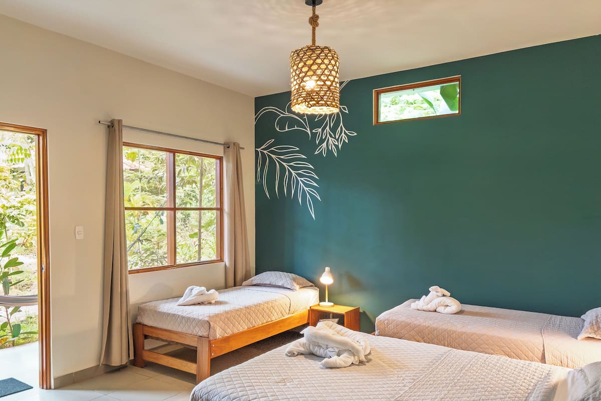 A room with two beds, a hanging light fixture, a window with curtains, and a wall with a leaf design.