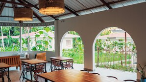 An indoor dining area with wooden tables and chairs, large windows, and hanging wicker lamps.