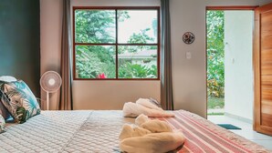 A bedroom with a bed, a window with curtains, a fan, and a view of greenery outside.