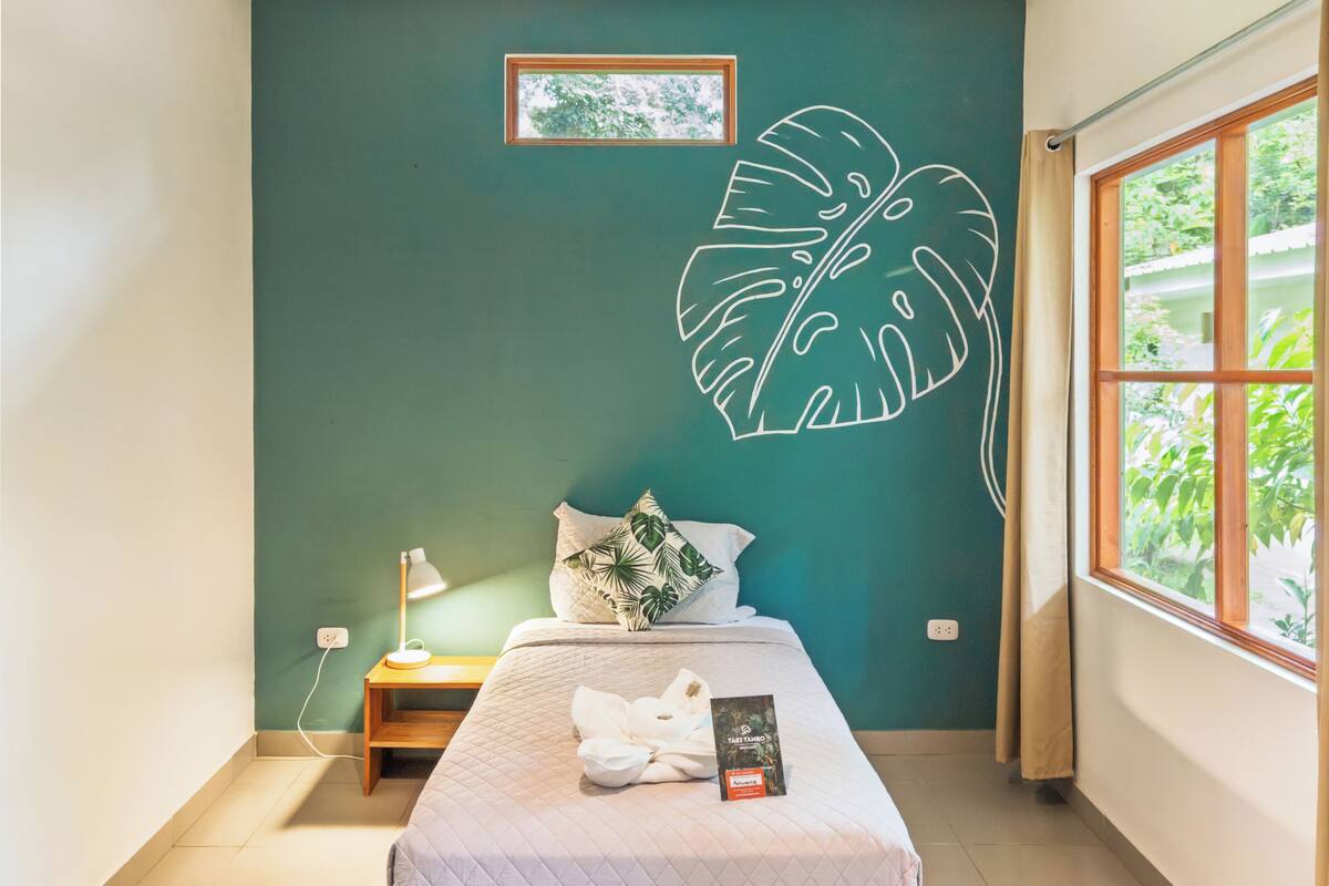 A bedroom with a bed, a nightstand, a lamp, a framed picture, and a large leaf mural on the wall.