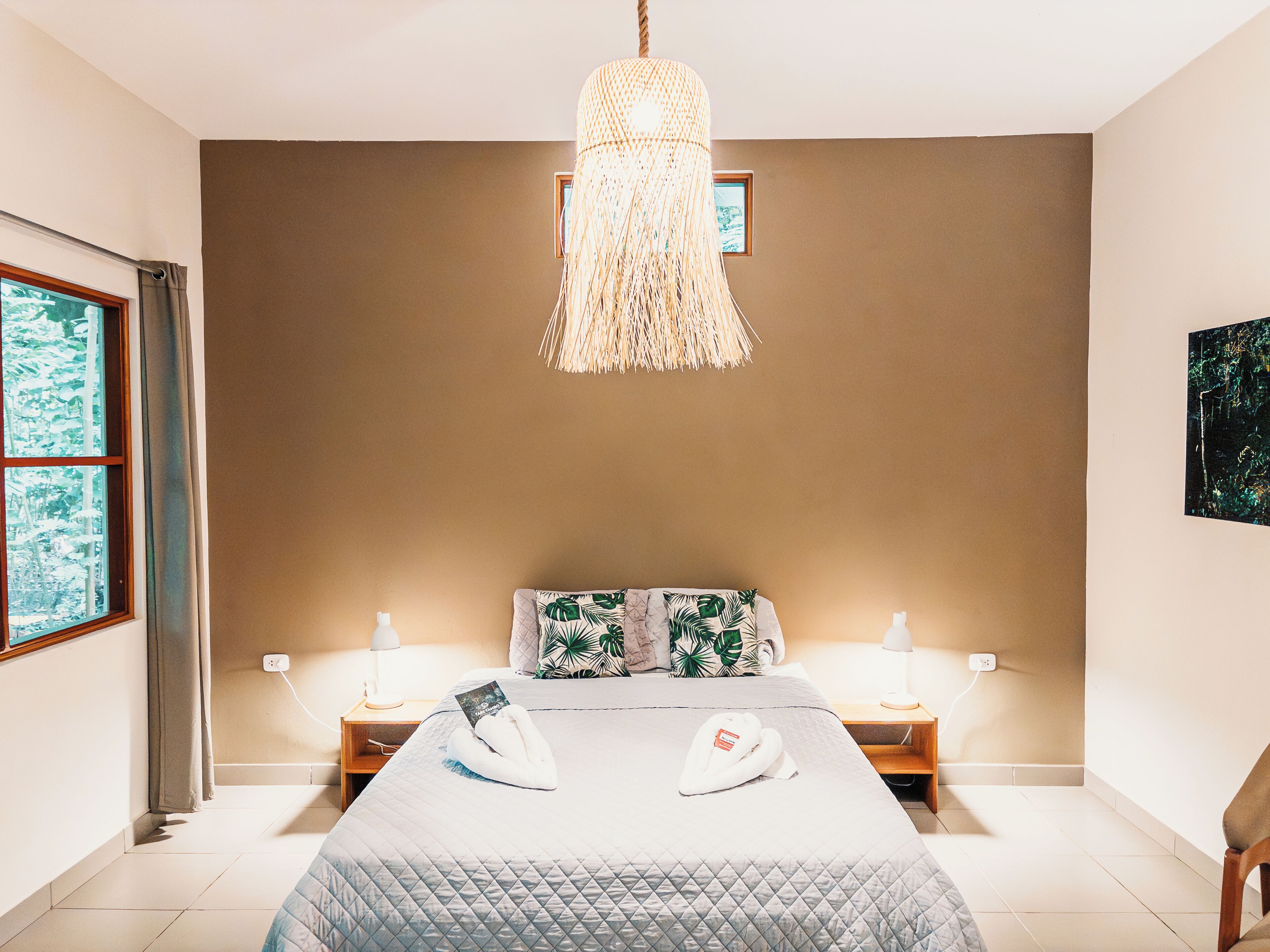 Un dormitorio con una cama, dos mesitas de noche, una lámpara colgante y una ventana con cortinas.