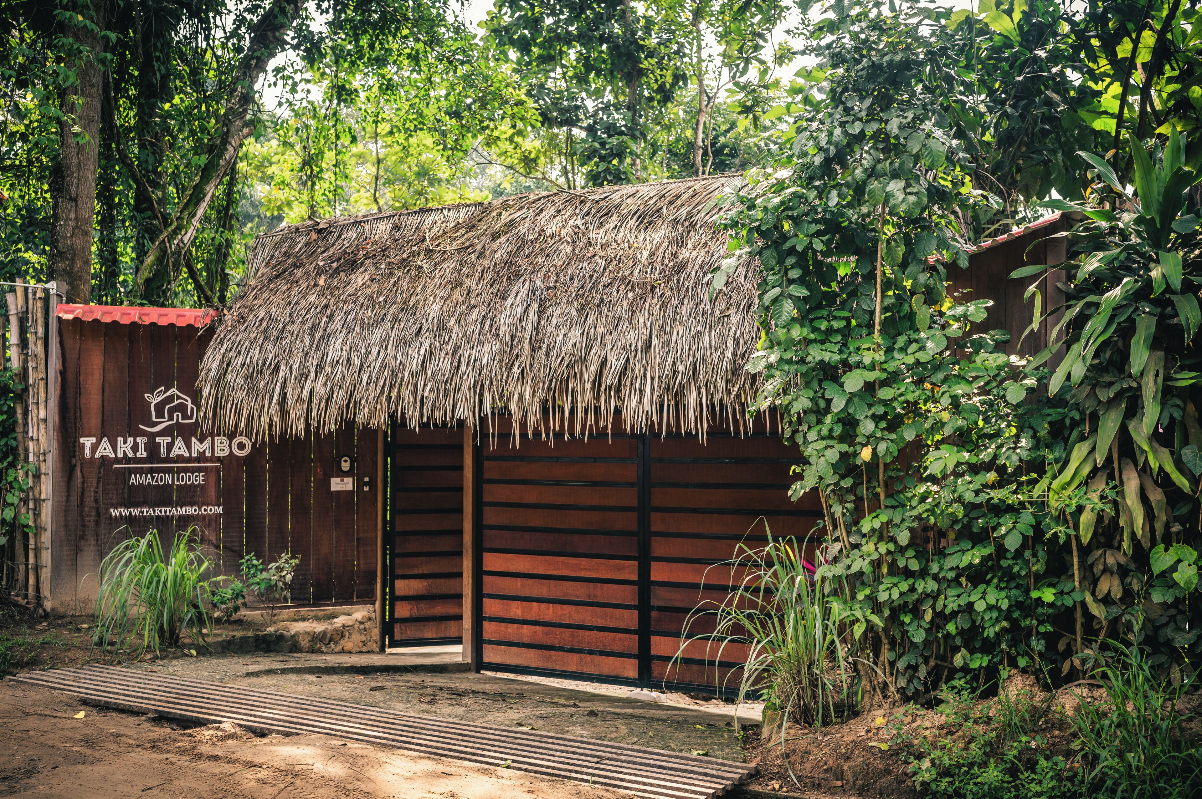 A rustic thatched-roof building with a wooden facade, labeled 'TAKI TAMBO AMAZON LODGE,' surrounded by dense greenery.