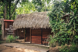 Una construcción rústica con techo de paja y fachada de madera, con la leyenda 'TAKI TAMBO AMAZON LODGE', rodeada de vegetación densa.