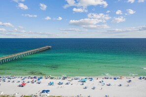 A beach with a pier extending into the sea, numerous beach umbrellas, and a clear sky.