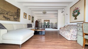 A spacious living room with a white sofa, a floral-patterned sofa, and a wooden coffee table. A dining area with green chairs and a kitchen is visible in the background.