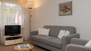 Un salon avec un canapé gris, un téléviseur posé sur un meuble en bois, une lampe sur pied et une petite table ronde sur laquelle sont posés des magazines.
