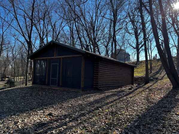 A log cabin with a large window and a door, surrounded by trees and a gravel path.