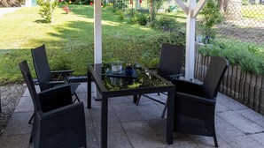 An outdoor patio with a table and four chairs, surrounded by a stone floor and a grassy area. - Guest at Gastel's FeWo 3-6 persons gladly workers or fitters (Fischach)
