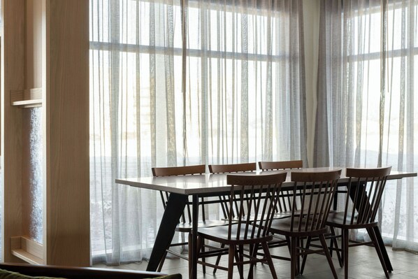 Un comedor con mesa y sillas, ventanas amplias con cortinas traslúcidas y piso de madera.
