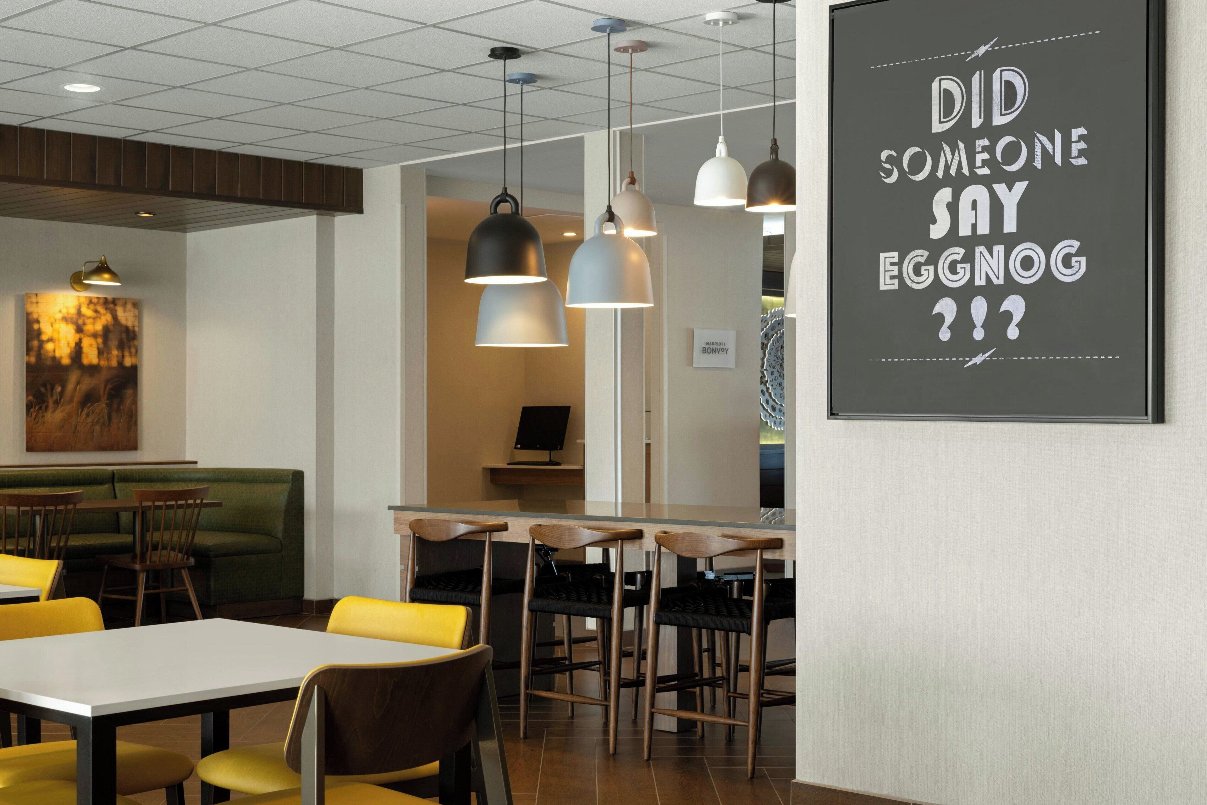 L’intérieur d’un restaurant moderne présente une salle à manger avec des chaises et des tables jaunes, un éclairage suspendu et un panneau mural avec du texte.