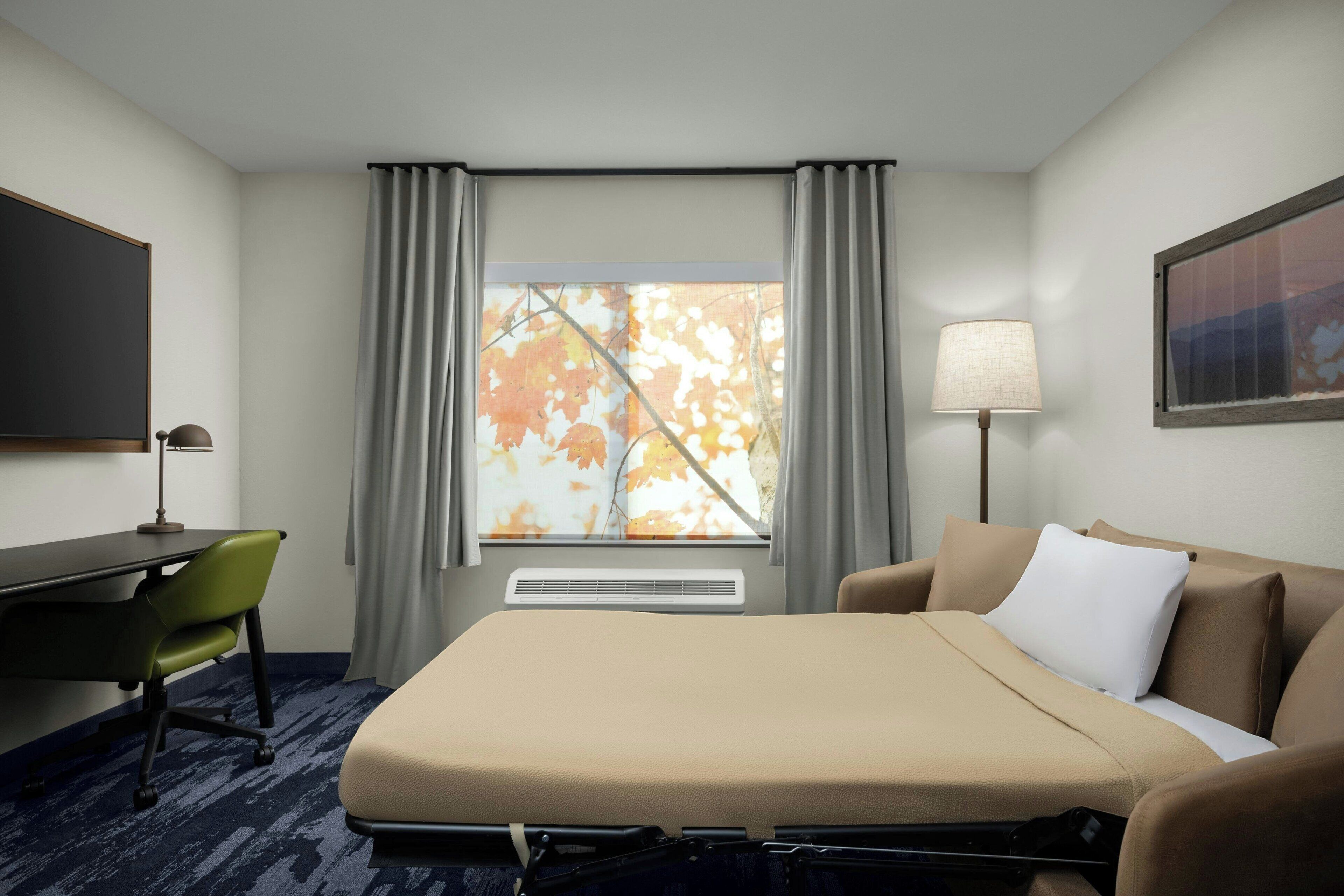 A hotel room with a bed, a desk, a chair, a lamp, and a window with curtains.