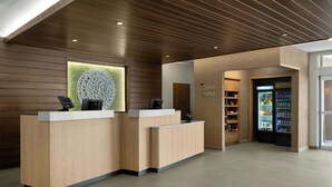 Un área de recepción con paneles de madera, un mostrador de recepción, un adorno mural y una tienda de ultramarinos en una esquina con estantes y una vitrina refrigerada.
