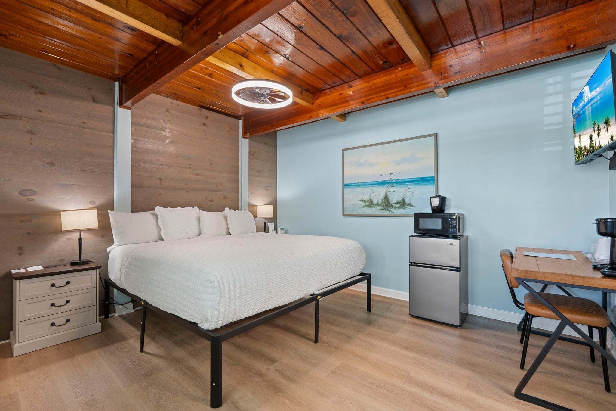 A bedroom with a bed, bedside tables, a desk, a chair, a refrigerator, and a painting on the wall.