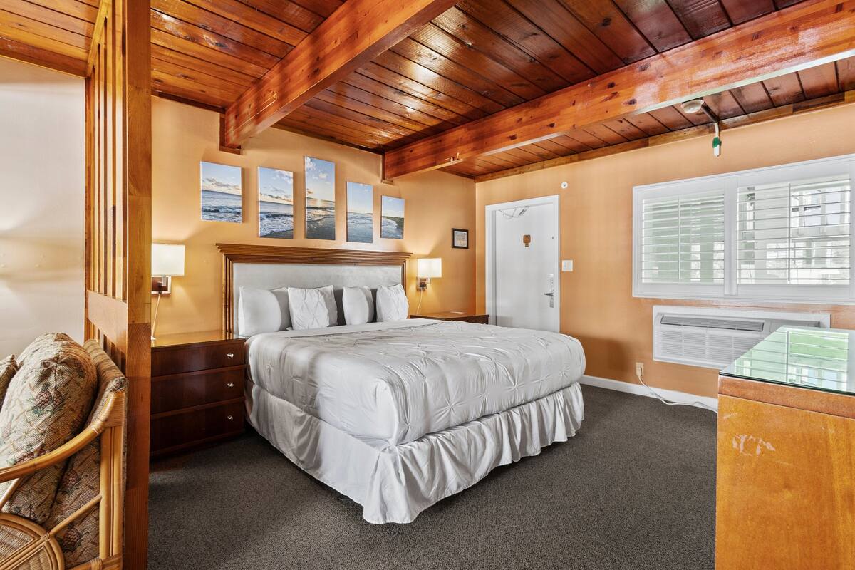 A bedroom with a wooden ceiling, a large bed with a white duvet, a bedside table with a lamp, and a window with blinds.