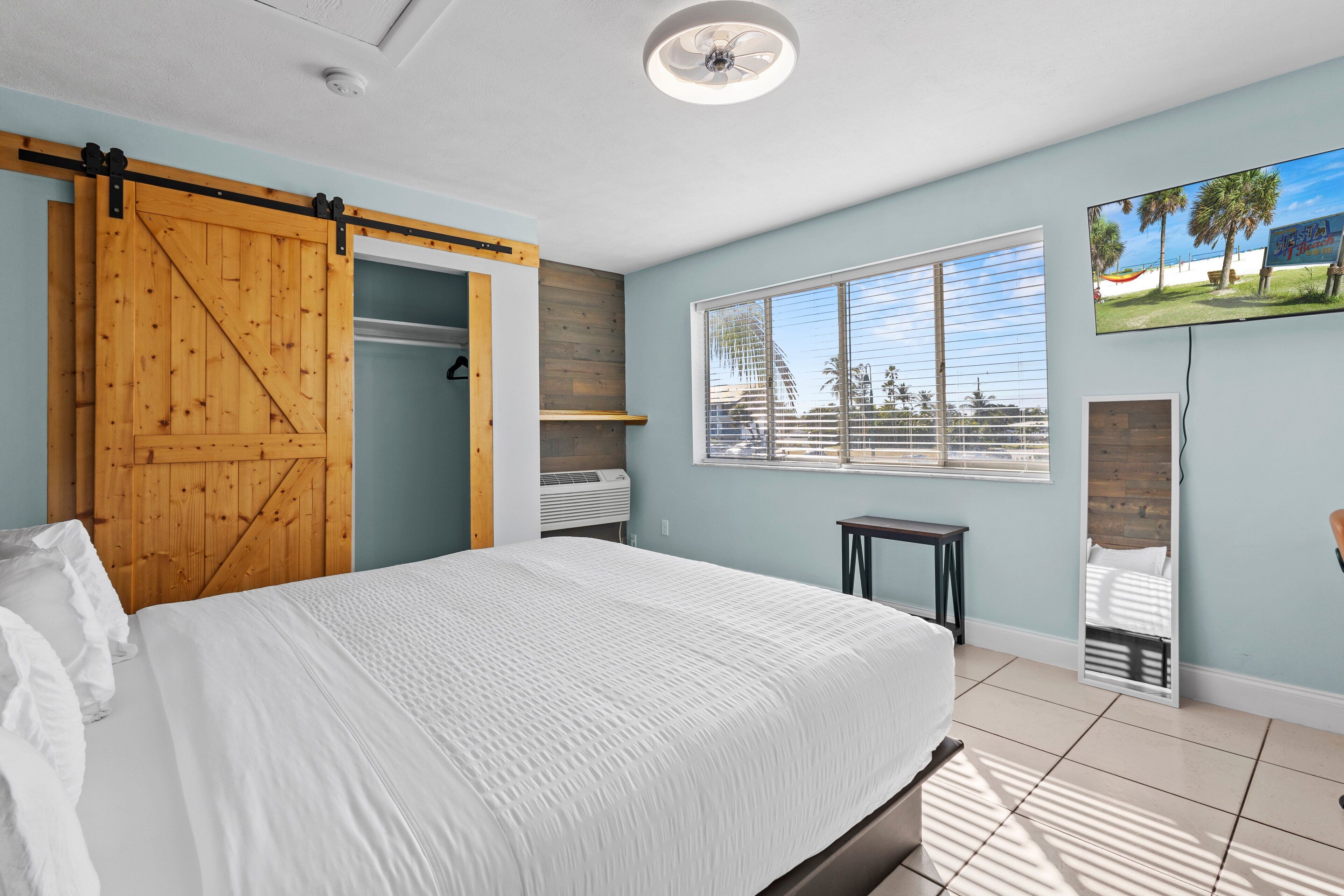 A bedroom with a large bed, a sliding barn door closet, a window with blinds, and a mounted TV.