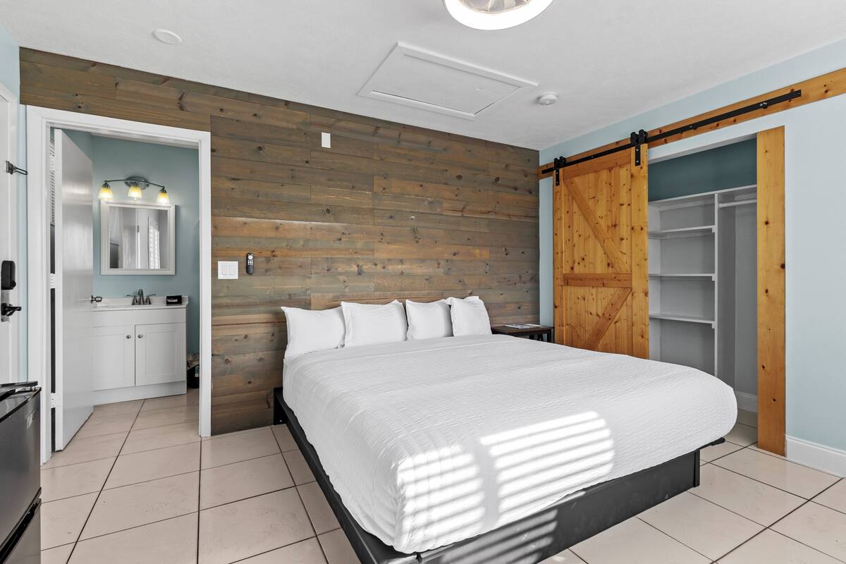 A bedroom with a bed, a wooden wall, a sliding closet door, and a bathroom with a mirror and sink.