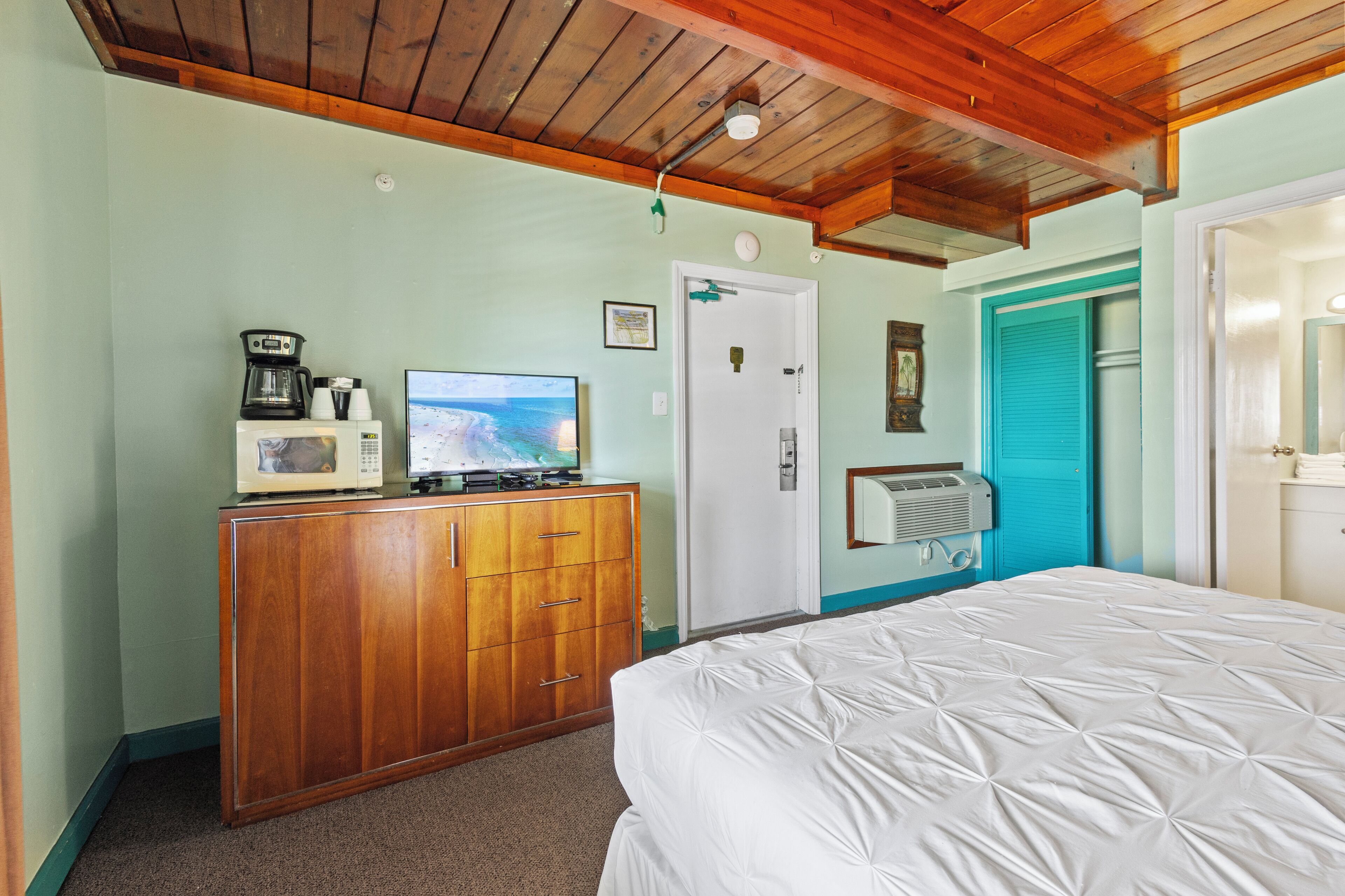 A bedroom with a wooden dresser, a bed, a television, and a door with a blue shutter.