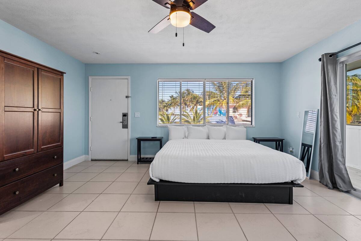 A bedroom with a bed, a wardrobe, a ceiling fan, a window with blinds, and a view of palm trees.