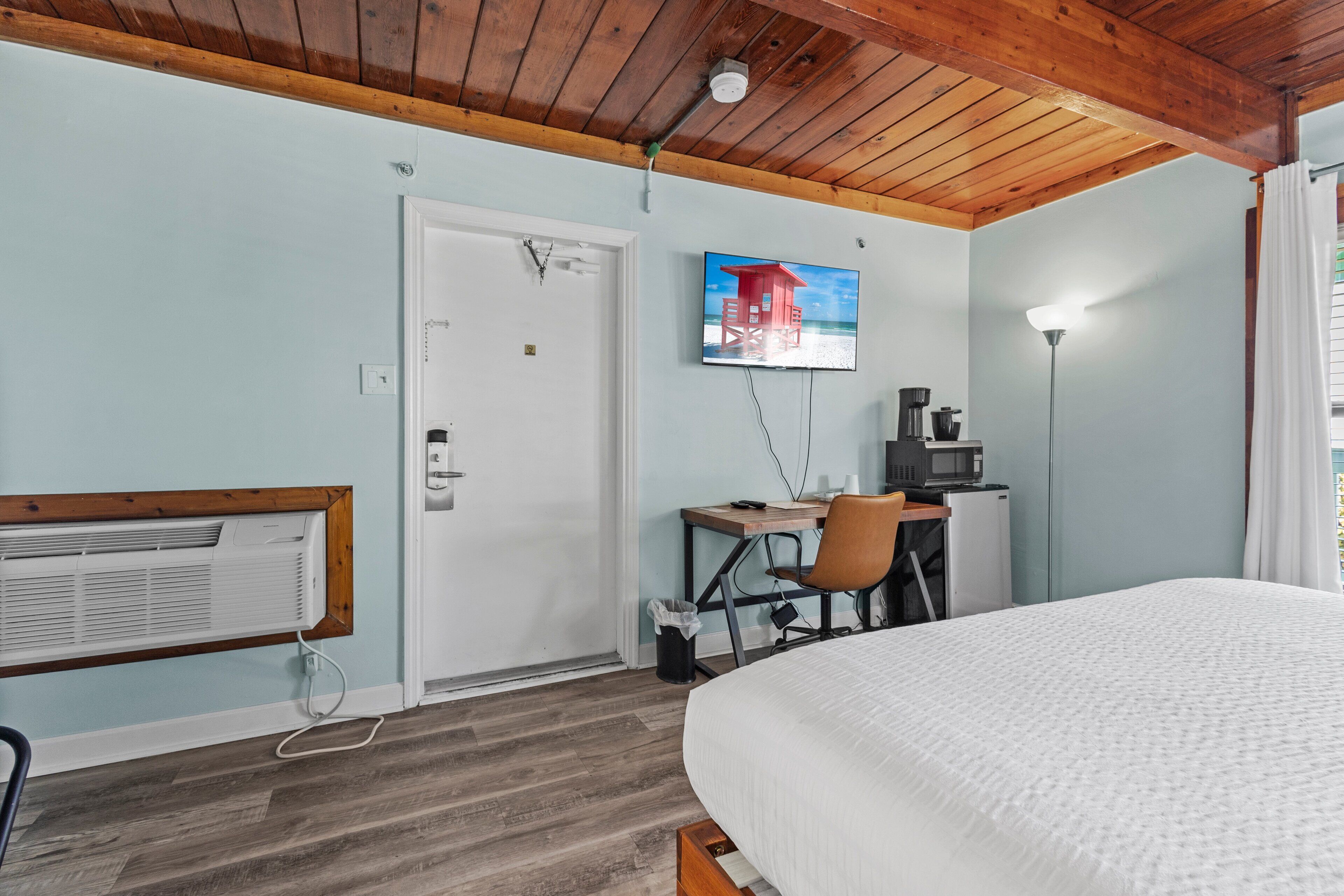 A hotel room with a wooden ceiling, a bed, a desk with a chair, a television, and a window with curtains.