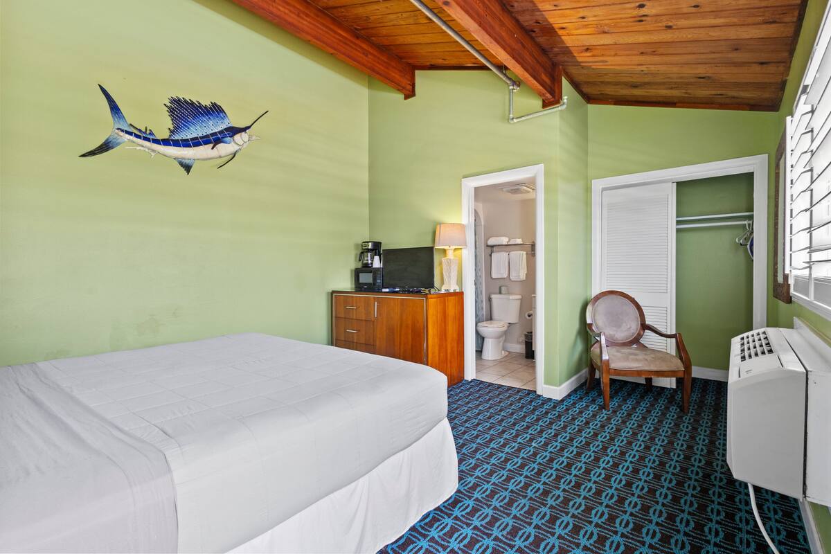 A room with a bed, a wooden dresser, a chair, a desk with a computer, and a wall-mounted lamp.