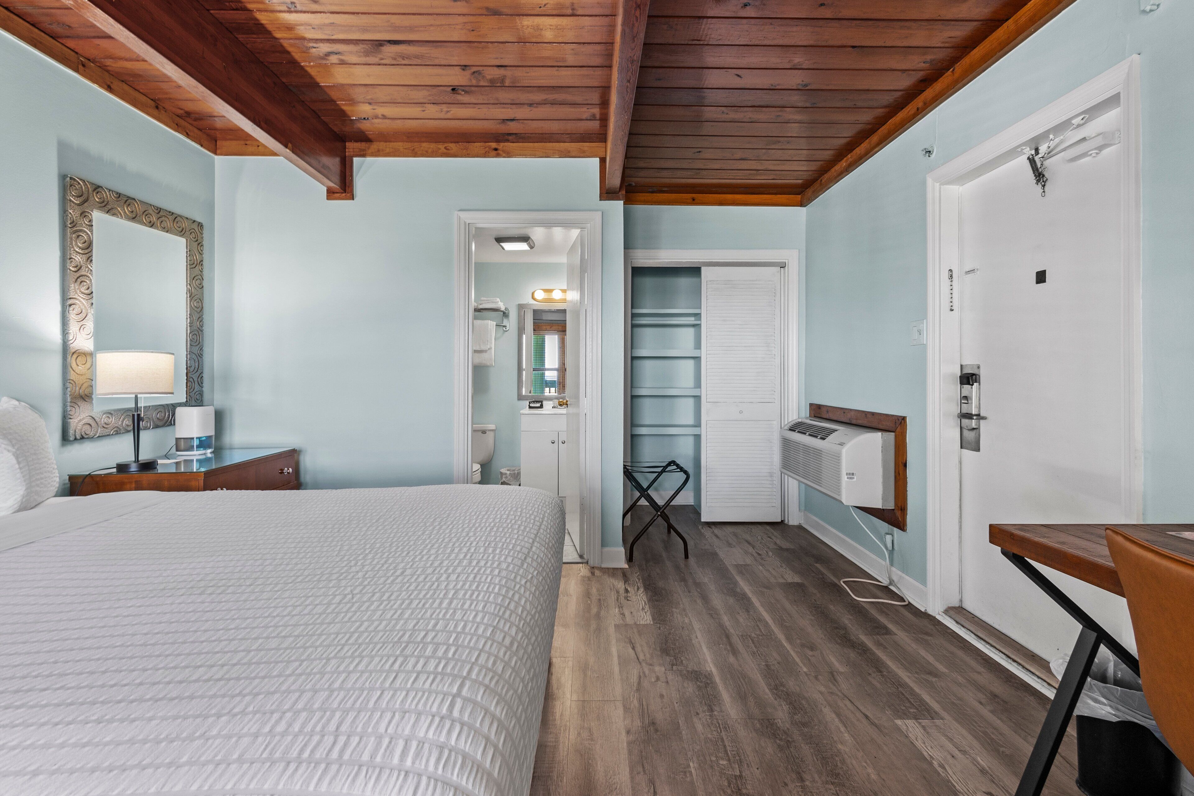 Een slaapkamer met een bed, nachtkastje en spiegel. Er is een deur naar een badkamer en een kledingkast.