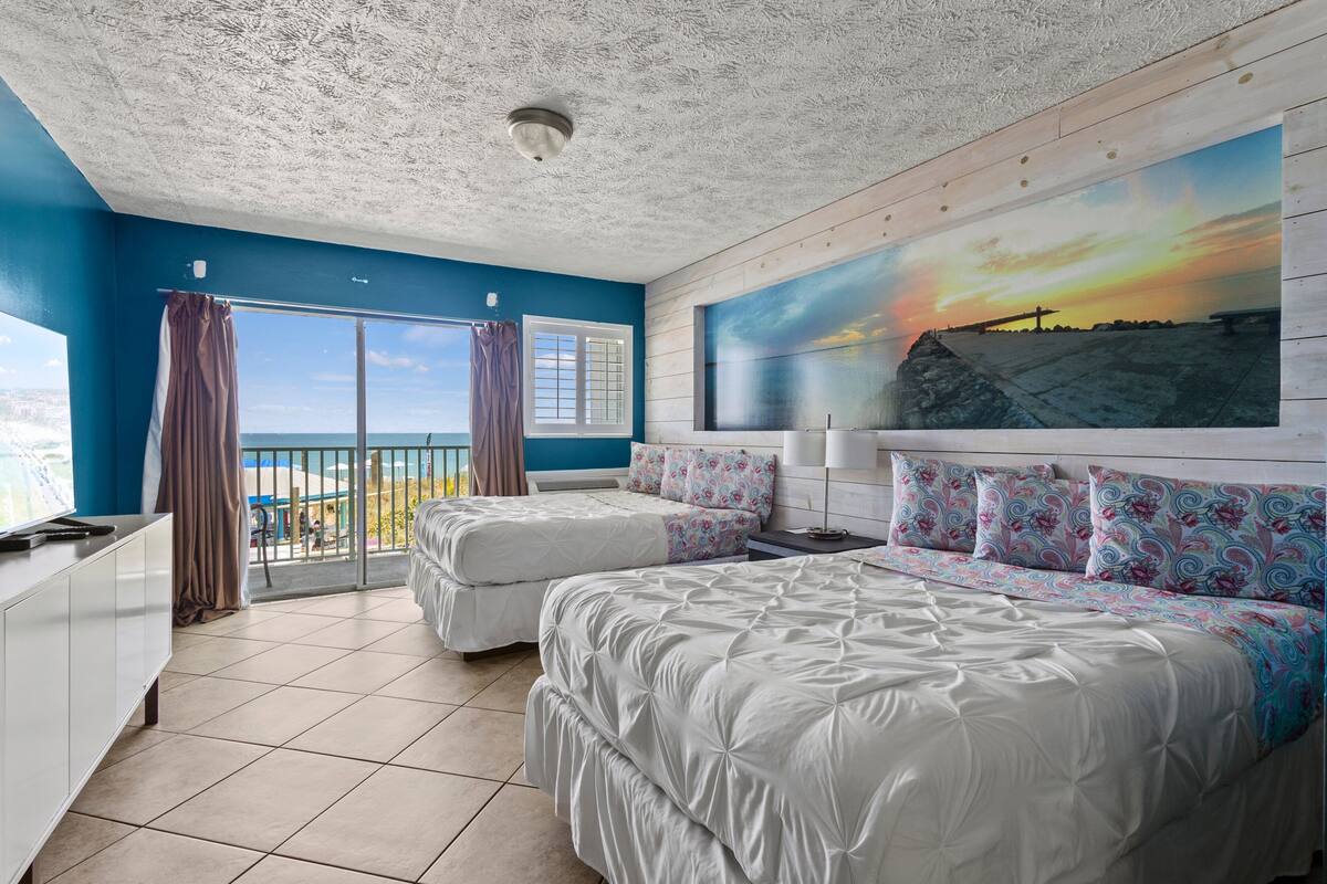 Une chambre avec deux lits, une grande fenêtre avec des rideaux, un tableau représentant un coucher de soleil et une vue sur l’océan.