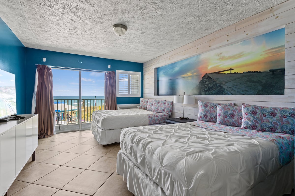 A bedroom with two beds, a large window with curtains, a painting of a sunset, and a view of the ocean.