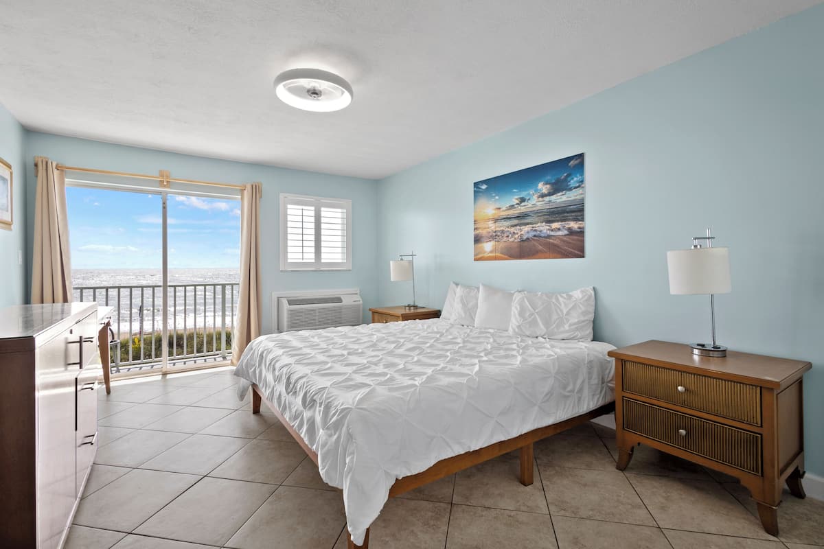 A bedroom with a bed, bedside tables, a dresser, and a view of the ocean through a balcony door.