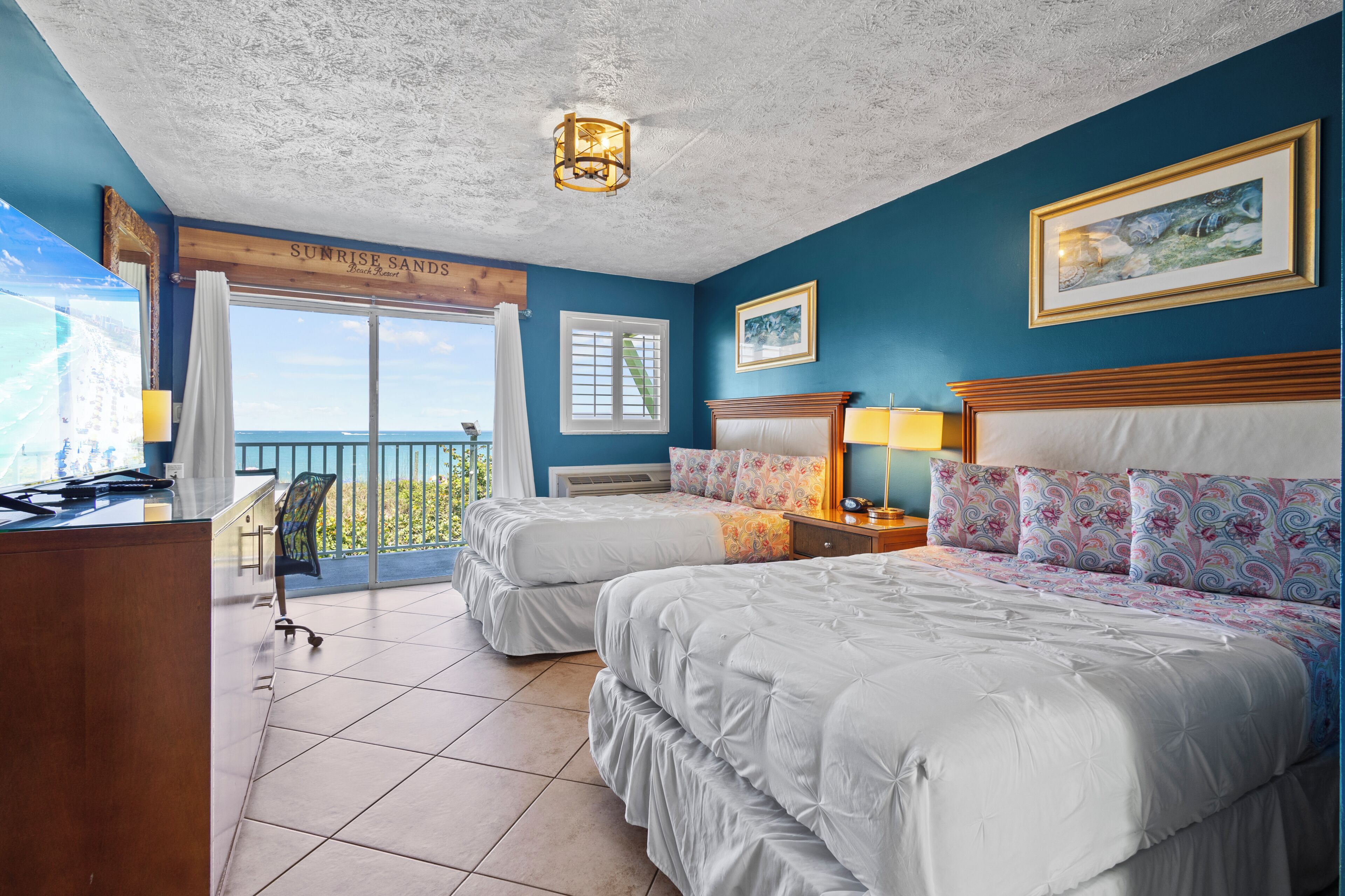 A hotel room with two beds, a desk, a chair, a balcony with a view, and a ceiling light fixture.