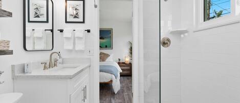 A modern bathroom with a shower, a sink, and a mirror. There is a bed visible through an open door in the background.
