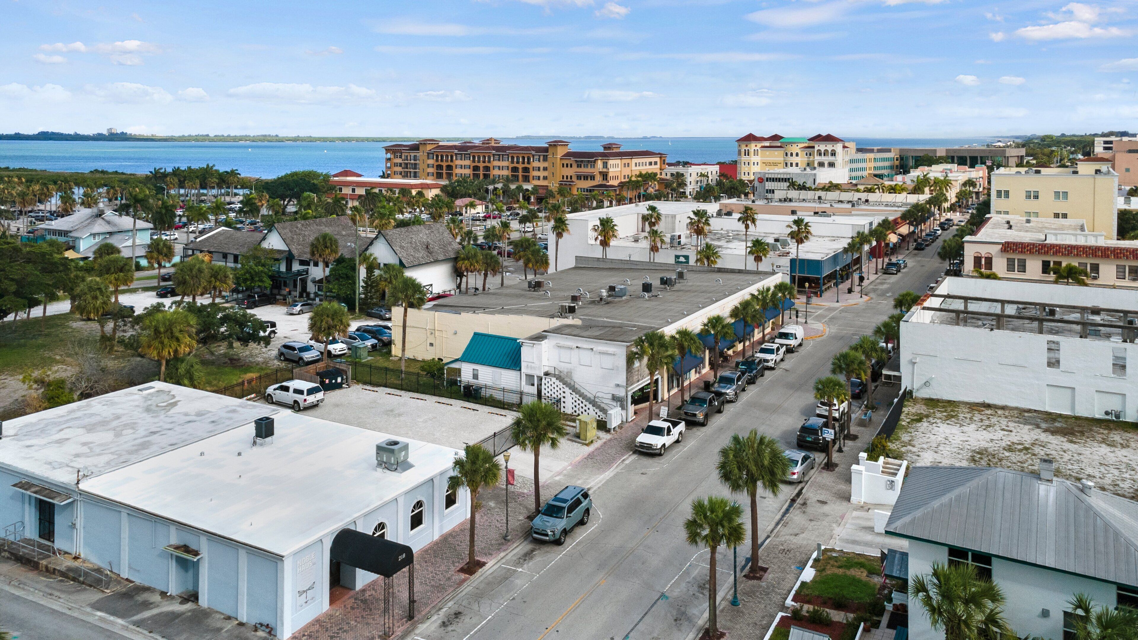 Un área urbana costera con una mezcla de edificios comerciales y residenciales, vista despejada al mar, y una calle bordeada de coches aparcados y palmeras.
