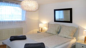Una cama bien tendida con edredón blanco y dos almohadas, una lámpara de noche, una mesita de noche pequeña y una lámpara colgante.