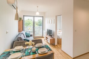 Een moderne, compacte woonruimte met een eethoek, een bank, een televisie en een balkon met uitzicht op groen.