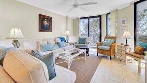 A spacious living room with a white ceiling fan, a large window with a view of trees, a beige sofa with blue cushions, and a wicker armchair with a cushion. - 123d Shipwatch by Avantstay Beach Access + Pool (Isle of Palms)