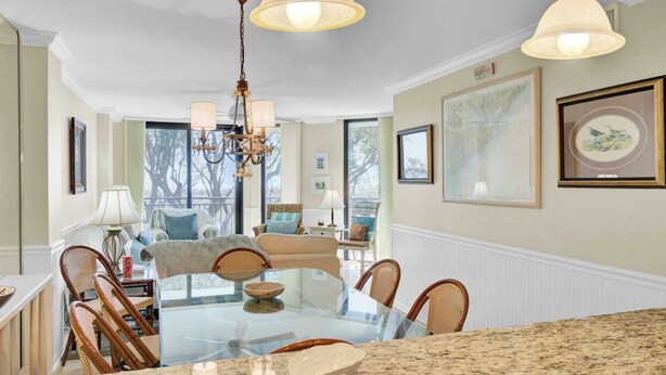 A dining area with a glass table, wicker chairs, and a chandelier. - 123d Shipwatch by Avantstay Beach Access + Pool (Isle of Palms)