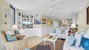 A spacious living room with a white sofa set, a wicker chair, a coffee table, and a television on a stand. - 123d Shipwatch by Avantstay Beach Access + Pool (Isle of Palms)