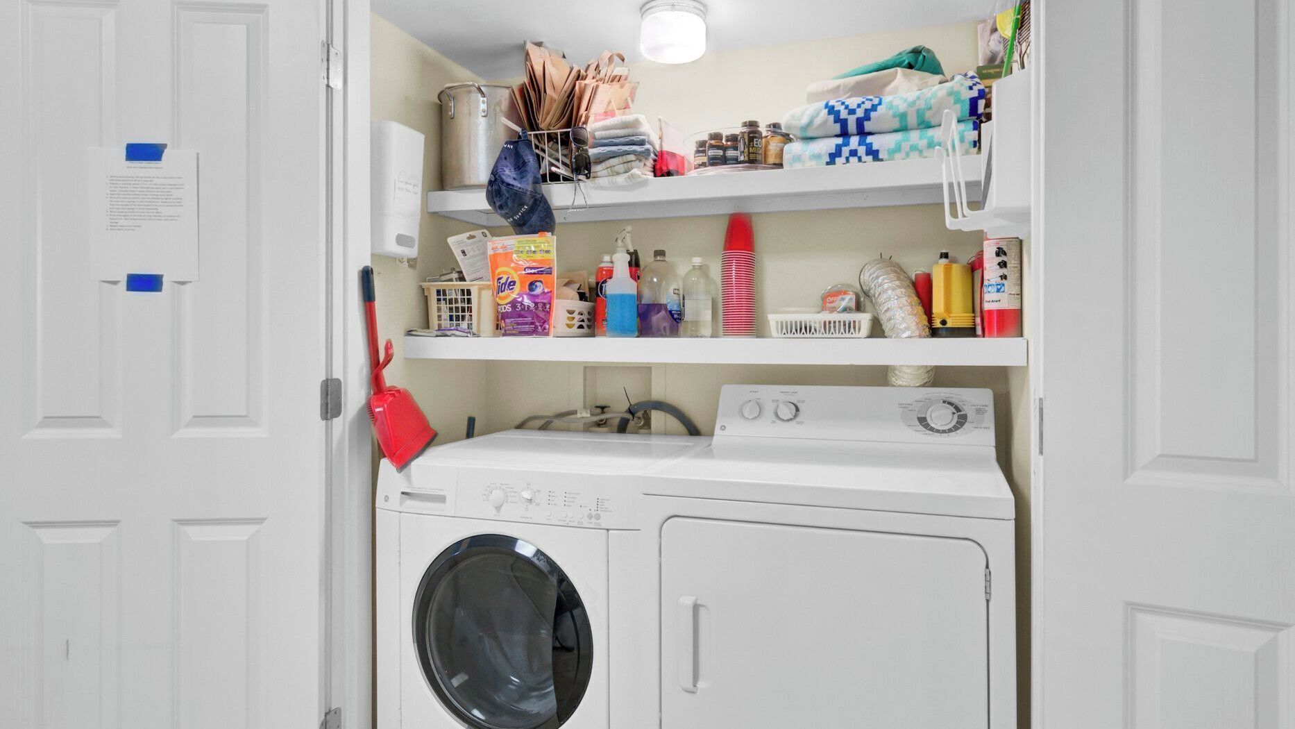 A laundry area with a washing machine, dryer, and shelves above containing cleaning supplies and folded clothes.