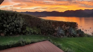 Ein Sonnenuntergang über einem See mit Bergen im Hintergrund, eine Holzterrasse und ein gepflegter Garten.