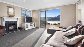 Ein Wohnzimmer mit Kamin, Fernseher und einer Couch mit Kissen. Große Fenster bieten Blick auf ein Gewässer und Berge.
