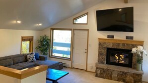 Ein geräumiges Wohnzimmer mit Kamin aus Stein, einem großen, an der Wand befestigten Fernseher und einer Glastür, die auf einen Balkon führt.
