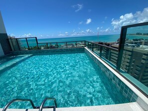 A rooftop infinity pool with clear blue water, surrounded by a glass railing, offering a view of the ocean and cityscape. - Smart Stay Apto 405 Mandi Hospitalidade (Maceió)