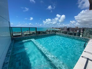 A rooftop infinity pool with clear blue water, surrounded by a glass balustrade, overlooking a cityscape and the ocean. - Smart Stay Apto 405 Mandi Hospitalidade (Maceió)
